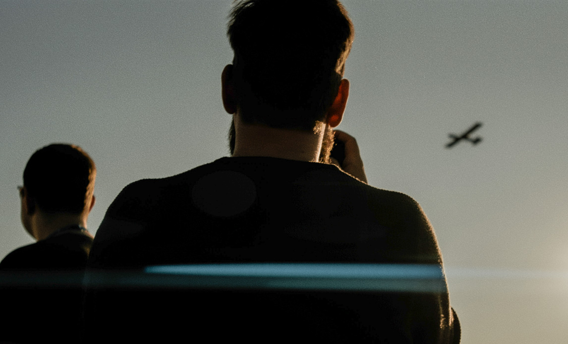 Siluetter af to mænd i forgrunden mens en drone flyver på himlen