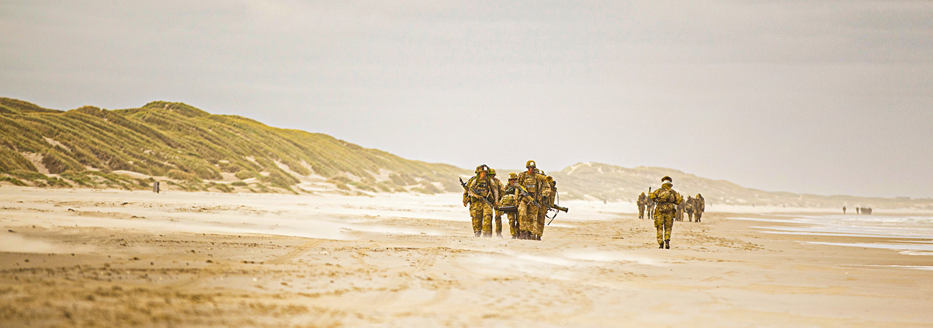 En grupper soldater på stranden under en øvelse. Fire soldater bærer den femte, som ligger på en båre.