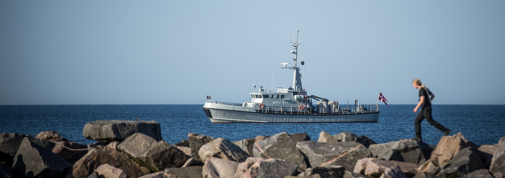 Militærfartøj passerer mole, hvor ung kvinde springer fra sten til sten