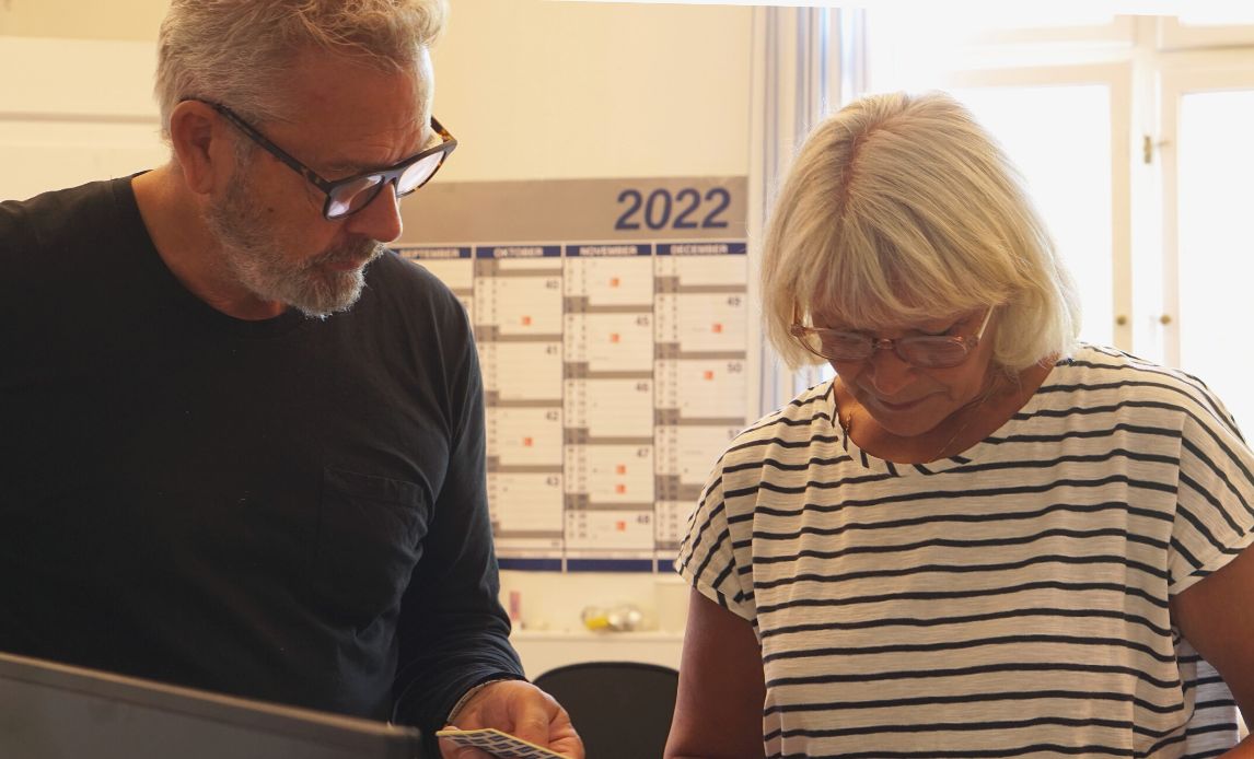 En mand (tv) og en kvinde (th) fra selektionssektionen står og drøfter dagens opgaver mens de kigger ned på deres noter. I baggrunden ses en kalender for 2022 og solen skinner ind ad vinduet.