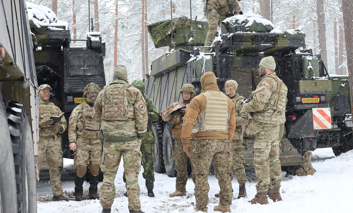 Otte soldater holder en briefing i en snebeklædt skov. De står i en cirkel, og rundt om dem er fire kampvogne. En soldat kravler på toppen af en af kampvognene.