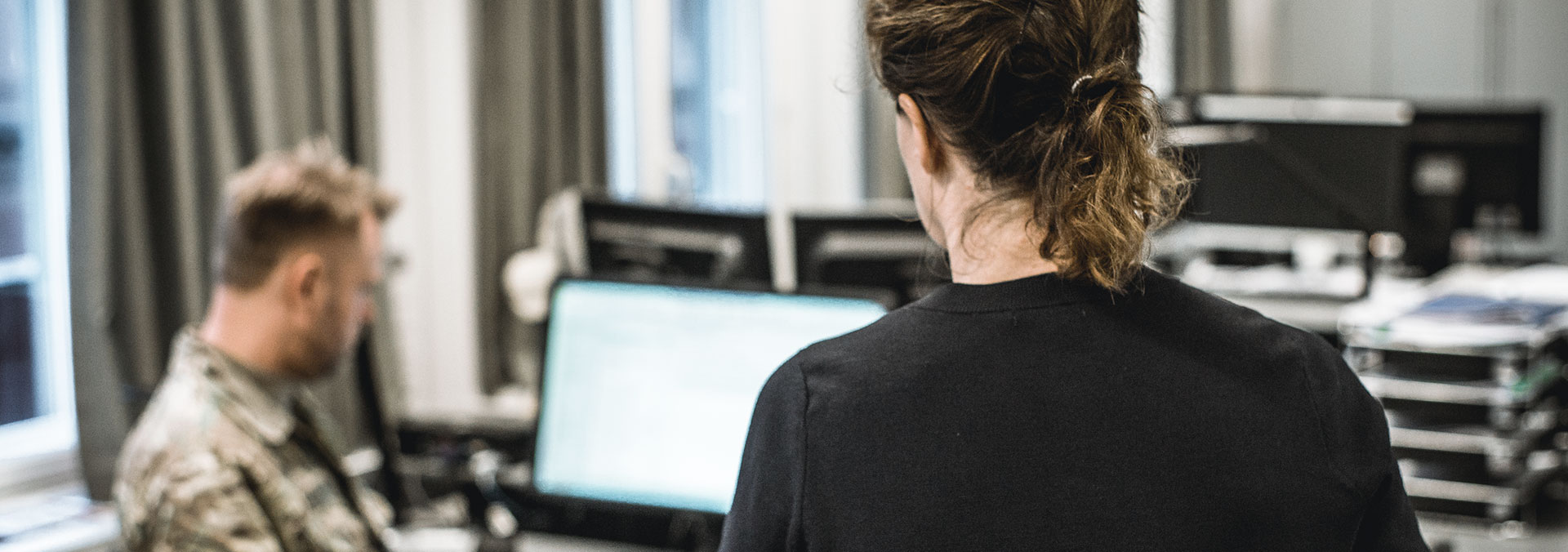 En kvindelig medarbejder står med ryggen til kameraet og kigger på en mandlig kollega i uniform, der sidder for sin computer