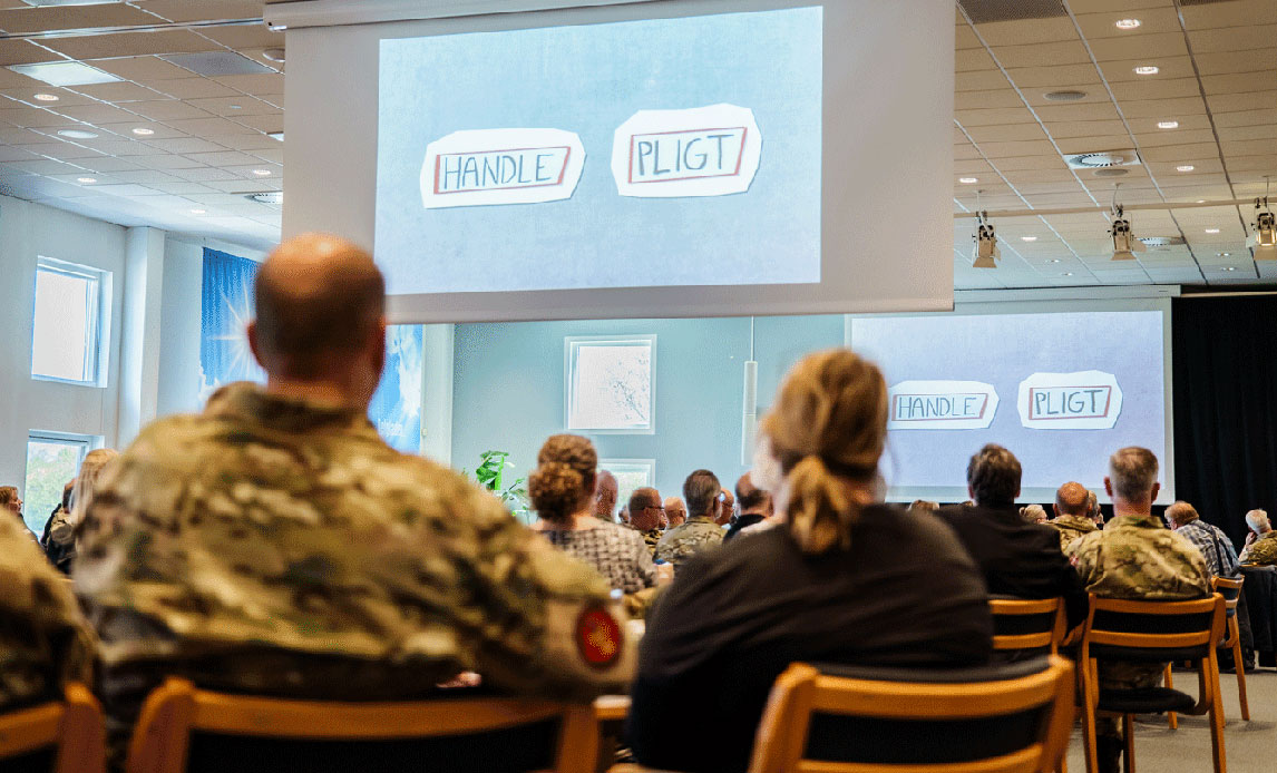 Billede fra en stor arbejdsmiljøkonference.
Deltagerne sidder med ryggen til kameraet. Nogle er i uniform. I baggrunden ses et lærred med en powerpointpræsentation, hvor der står handlepligt.