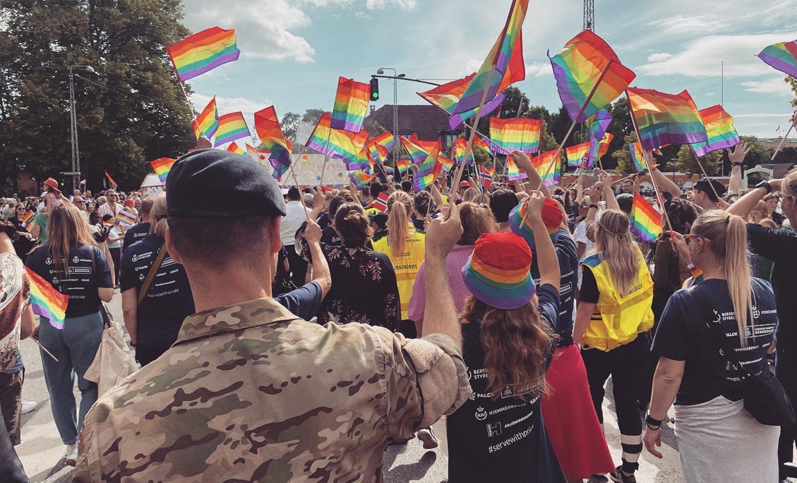 Billede fra pride-paraden 2022 i København. Billedet er tage en anelse oppefra, og personerne på billedet har alle ryggen til. Forrest i billedet ses en mand i uniform. Resten er primært civile deltagere i ens mørkeblå t-shirts med logoer på ryggen. Alle deltagere flager med små regnbueflag.