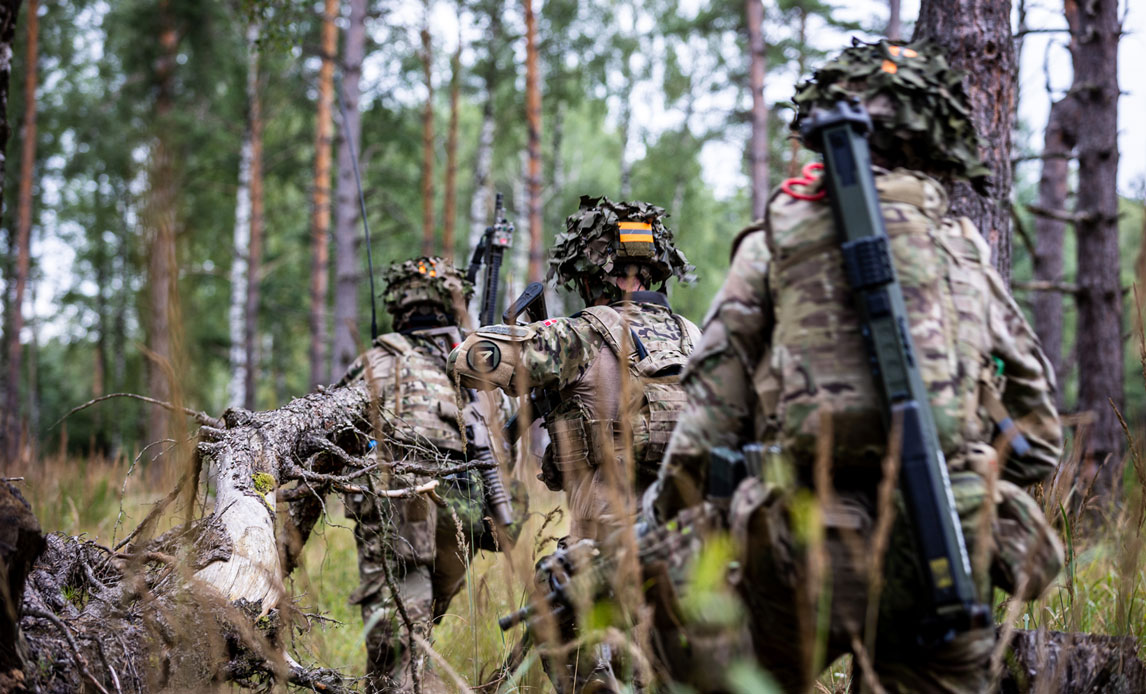 Tre soldater går i en skov med ryggen til kameraet. De bærer hjelm og våben.